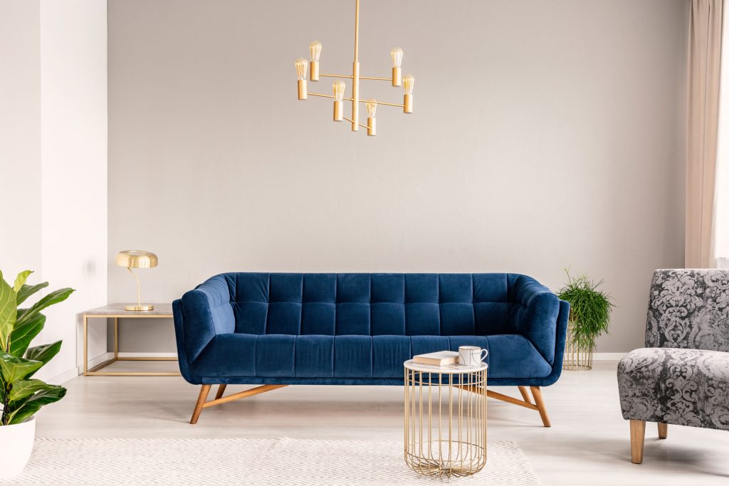 Gold lamp hanging above royal blue sofa in real photo of light grey sitting room interior with empty wall